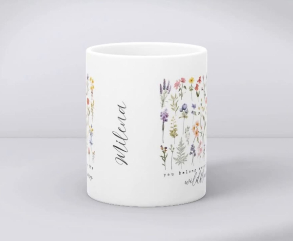 Detailansicht einer weißen Keramiktasse mit Wildblumenmotiv, dem Zitat „You belong among the wildflowers“ und dem personalisierten Namen „Milena“ in eleganter Schrift.